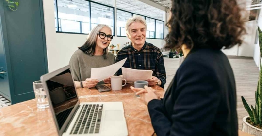 An older man and woman holding paper while another woman with a laptop sits across from them